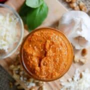 A jar of red pepper pesto sauce sits on a wooden board, surrounded by garlic, grated cheese, basil leaves, walnuts, and a tomato.