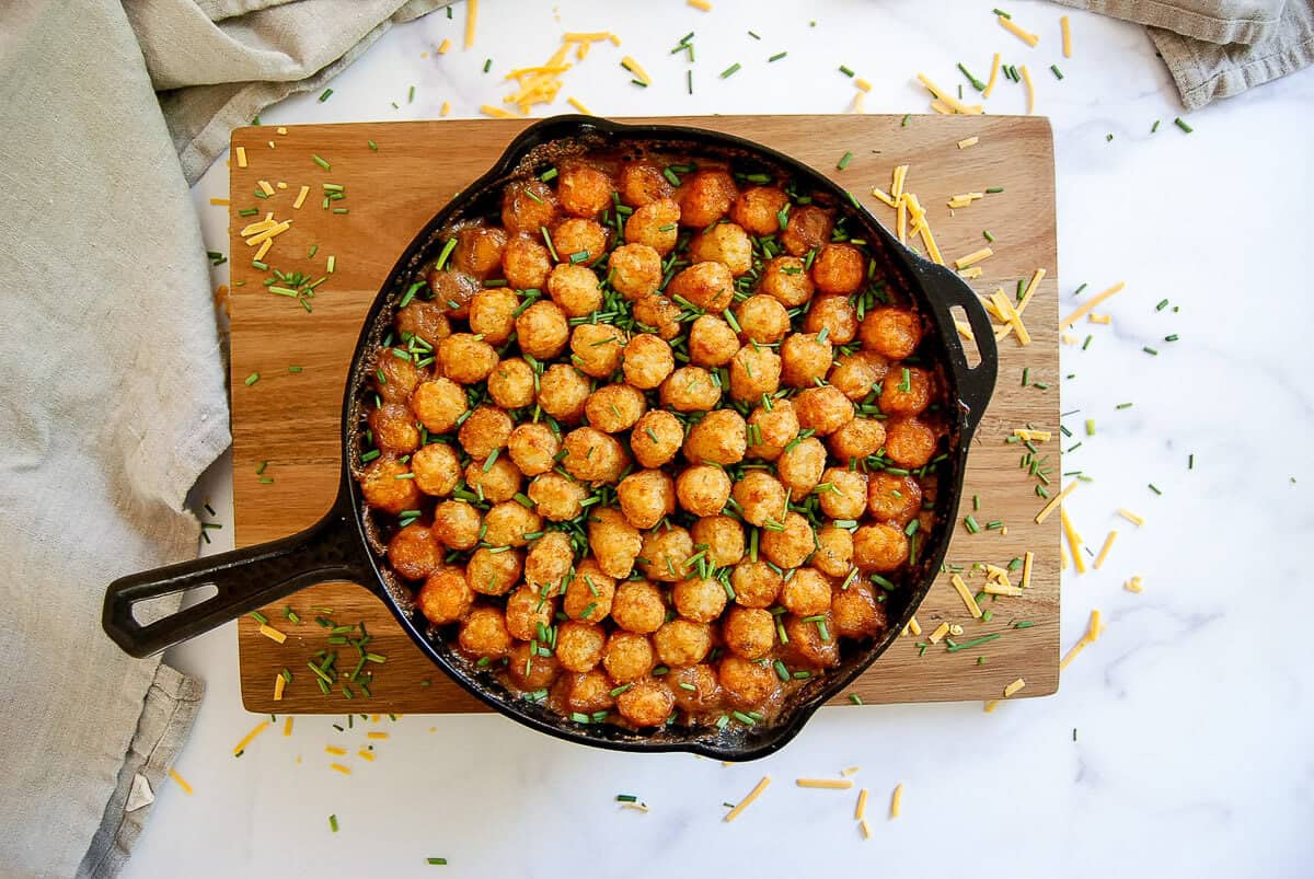 A cast-iron skillet sits on a wooden board, filled with tater tot casserole topped with golden tater tots and chopped chives, with cheese and chives scattered around.
