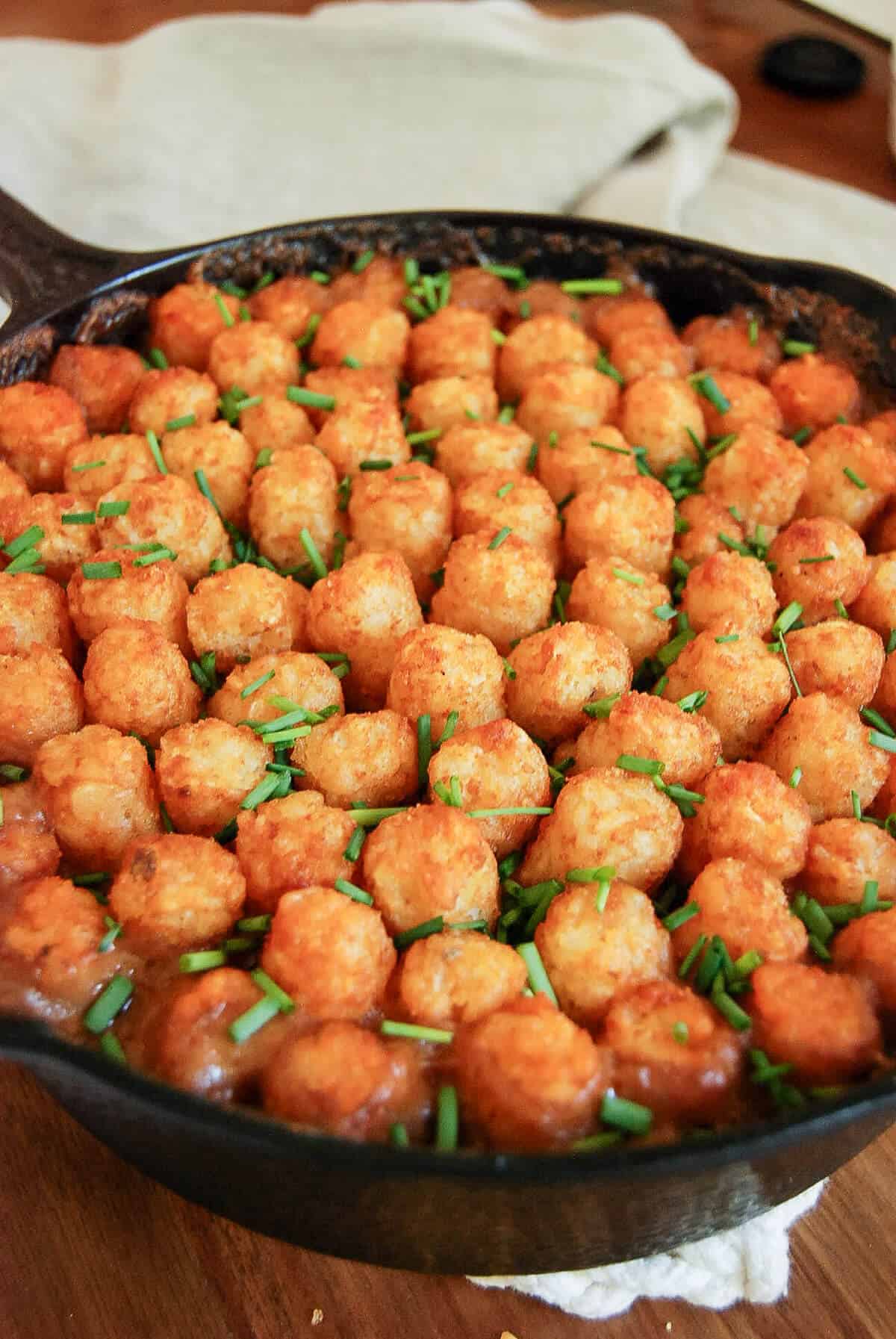A cast iron skillet filled with tater tot casserole, topped with rows of golden tater tots and sprinkled with chopped chives, sits on a wooden table.