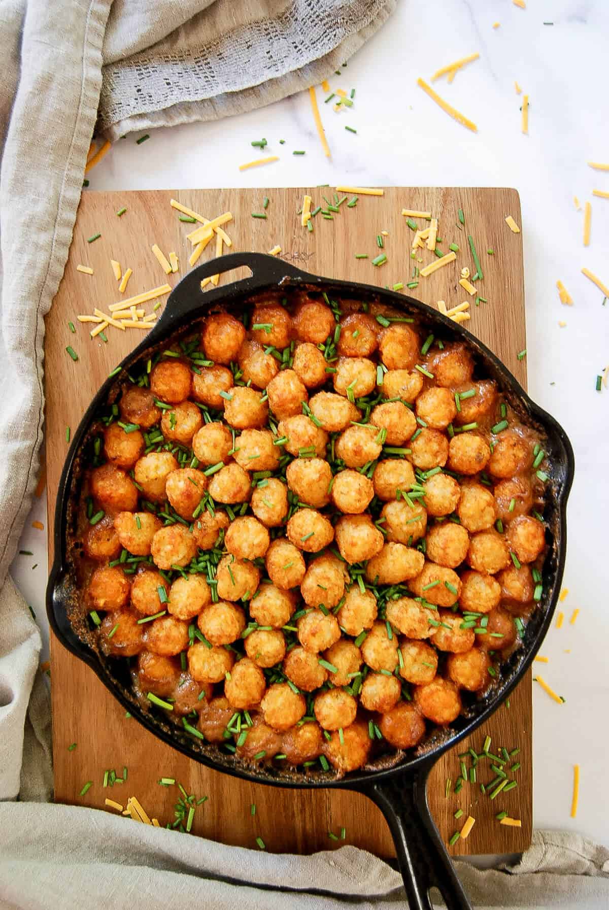 A cast iron skillet filled with tater tot casserole, topped with golden tater tots and sprinkled with chopped chives, sits on a wooden board with some shredded cheese scattered around.