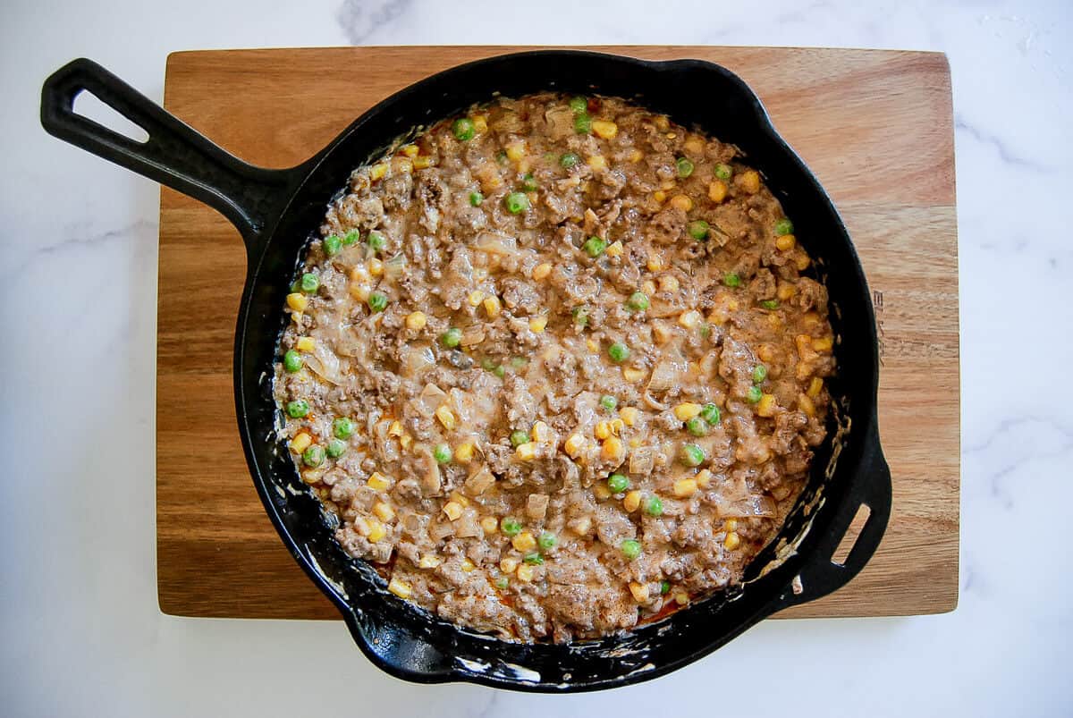 A cast iron skillet filled with a creamy casserole mixture of ground beef, peas and corn sits on a wooden cutting board atop a white countertop.