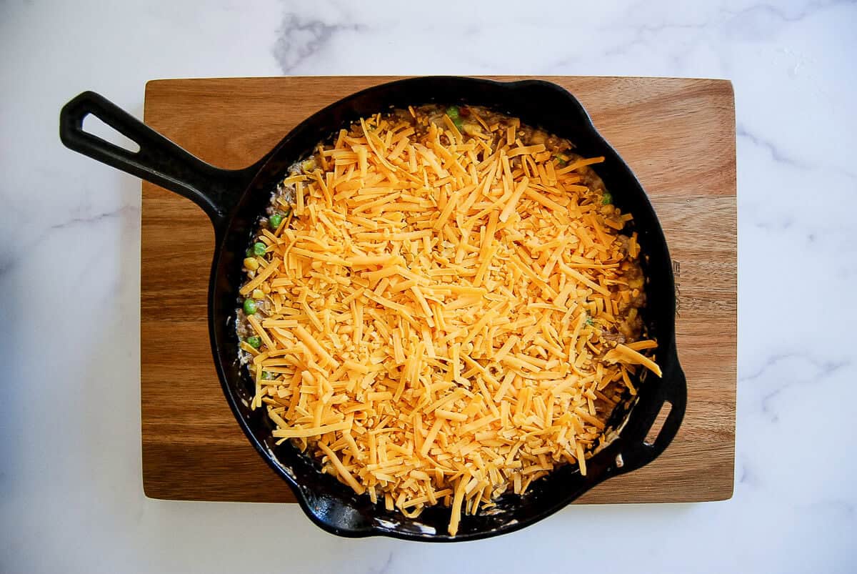 A cast iron skillet filled with a casserole topped with shredded cheddar cheese sits on a wooden cutting board atop a white marble surface.