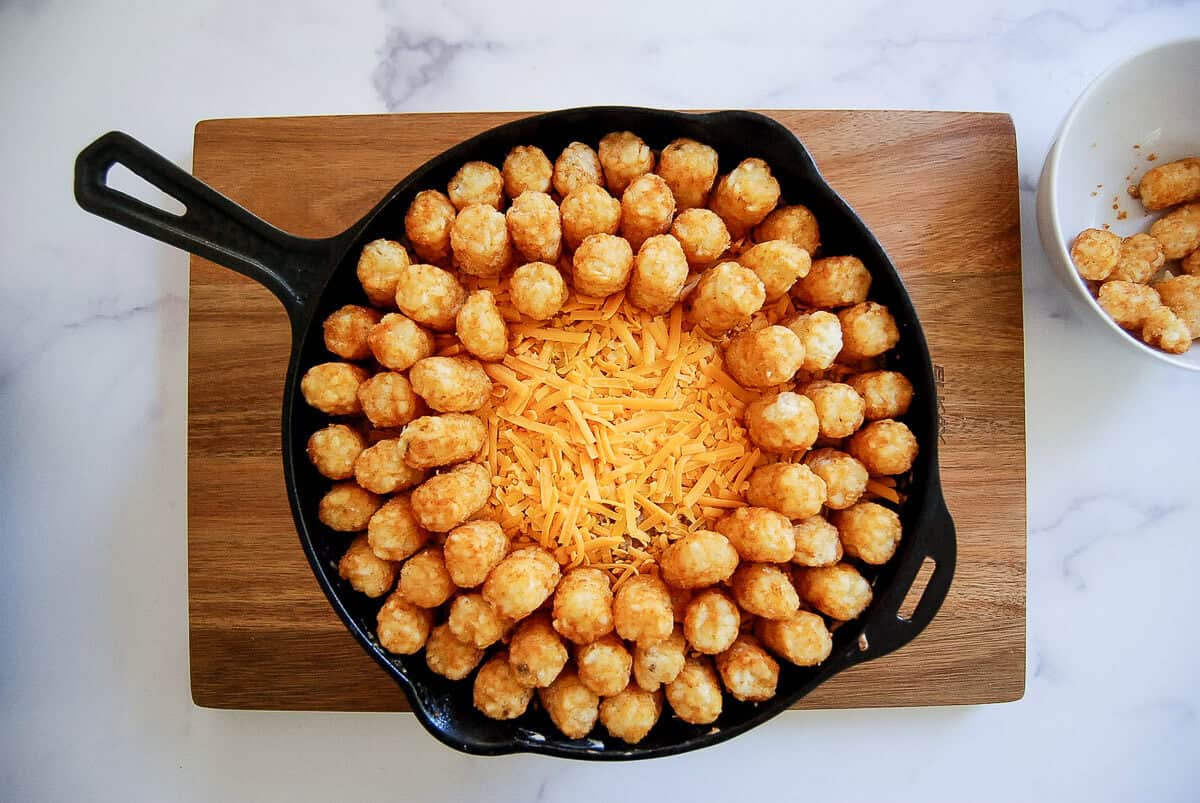 A cast iron skillet on a wooden board filled with shredded cheddar cheese in the center and tater tots arranged in a ring around the edge. A white bowl with a few tater tots is beside the skillet.