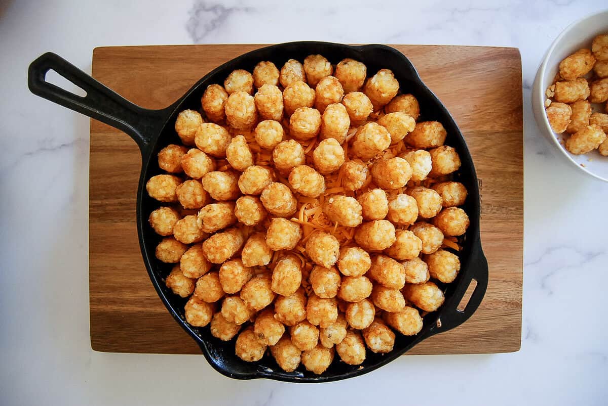 A cast iron skillet filled with neatly arranged tater tots sits on a wooden cutting board, with a small bowl of tater tots visible to the right.