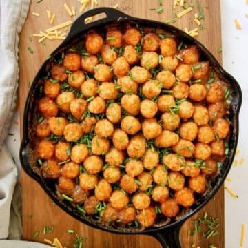 A cast iron skillet filled with a tater tot casserole, topped with rows of golden tater tots and garnished with chopped chives, sits on a wooden board.