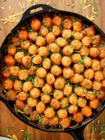 A cast iron skillet filled with a tater tot casserole, topped with rows of golden tater tots and garnished with chopped chives, sits on a wooden board.