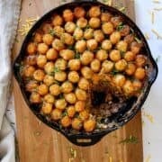 A cast iron skillet filled with a casserole topped with golden tater tots and chopped chives, with a serving scooped out, on a wooden board with shredded cheese scattered around.