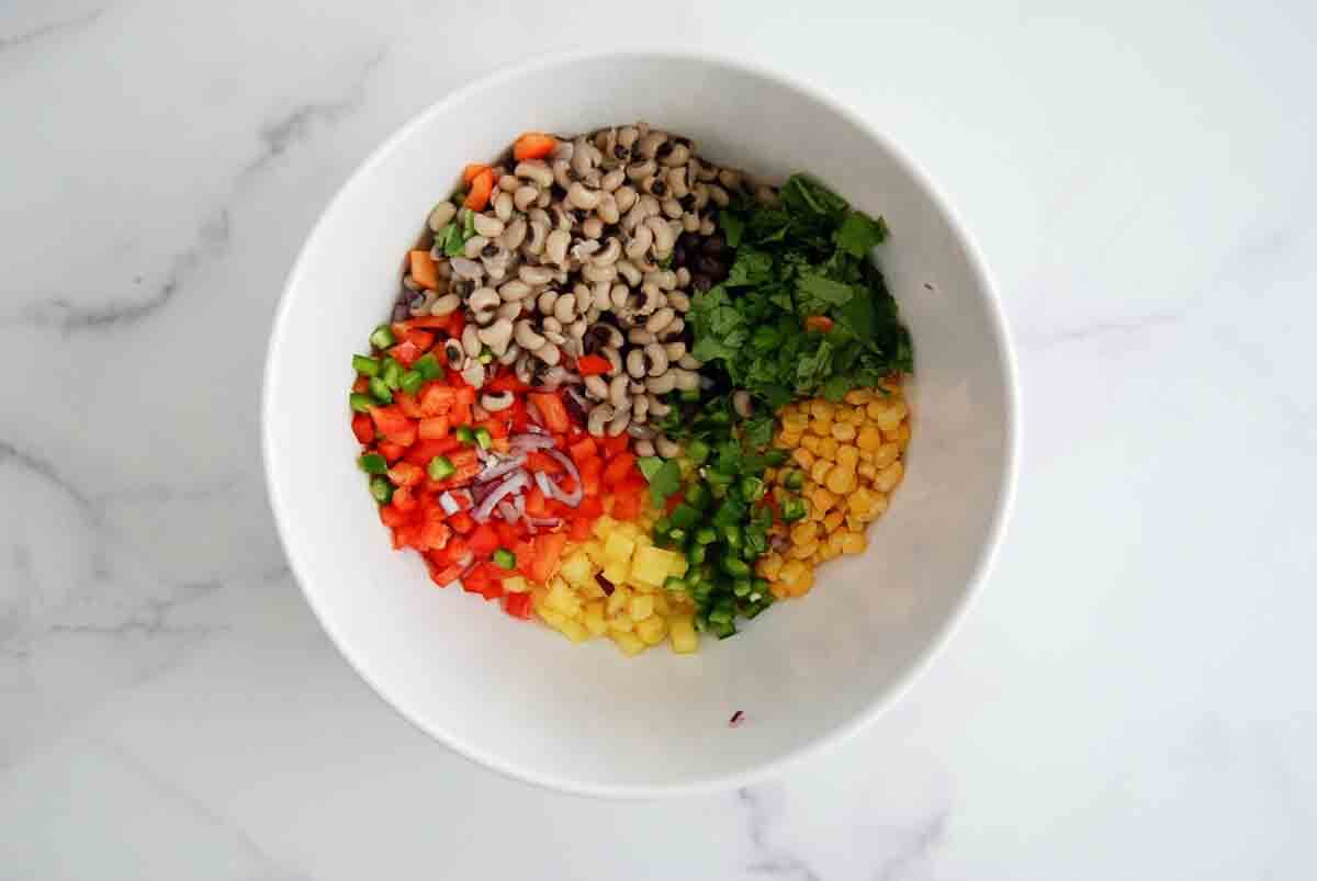 A white bowl with neatly separated ingredients: black-eyed peas, chopped cilantro, corn, diced red bell pepper, diced orange bell pepper, jalapeño, pineapple, and thinly sliced red onion on a marble surface.