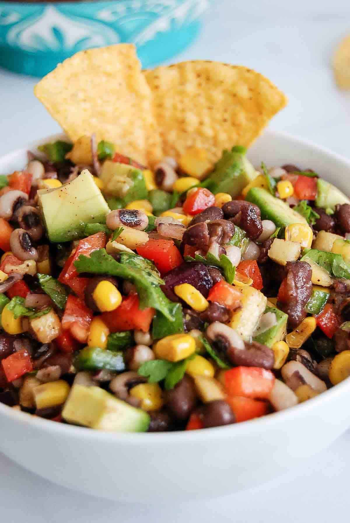 A bowl of cowboy caviar salad with black beans, corn, tomatoes, avocado, and cilantro, garnished with two tortilla chips.