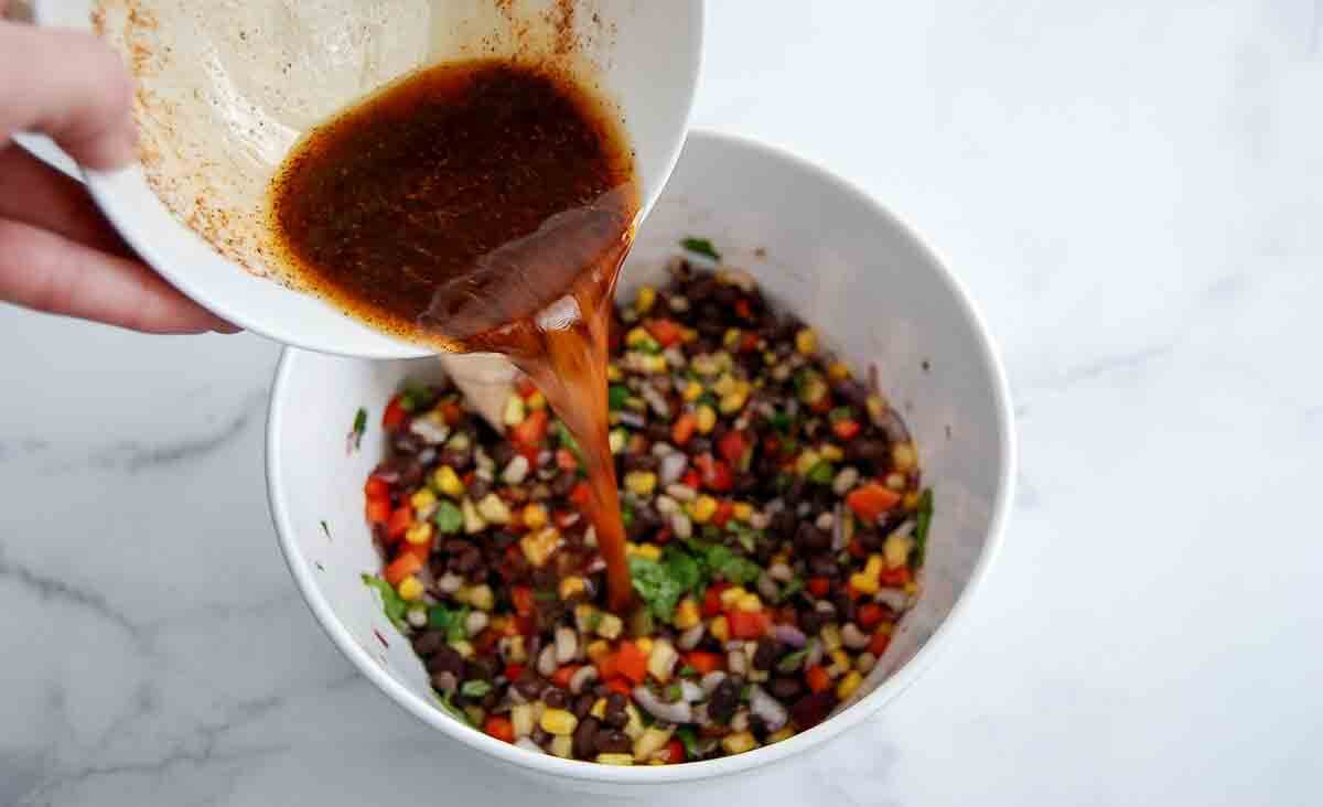 A hand pours dressing from a bowl into a white bowl filled with black bean salad, including chopped peppers, onions, and corn for a cowboy caviar dip.