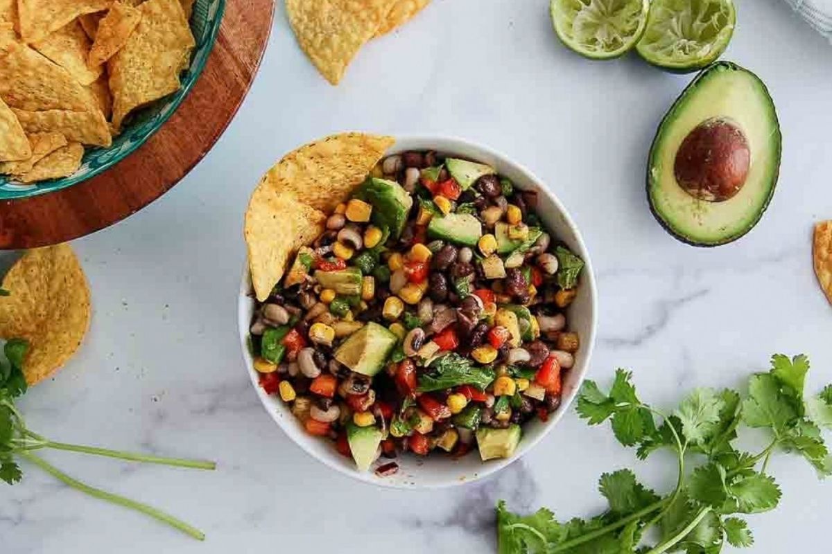 Bowl of texas caviar with diced avocado, surrounded by tortilla chips, lime halves, cilantro, and a halved avocado on a white surface.