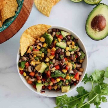 A bowl of texas caviar dip with beans and vegetables, avocado, corn, and peppers, garnished with a tortilla chip. Fresh avocado, cilantro, lime, and more chips are nearby.