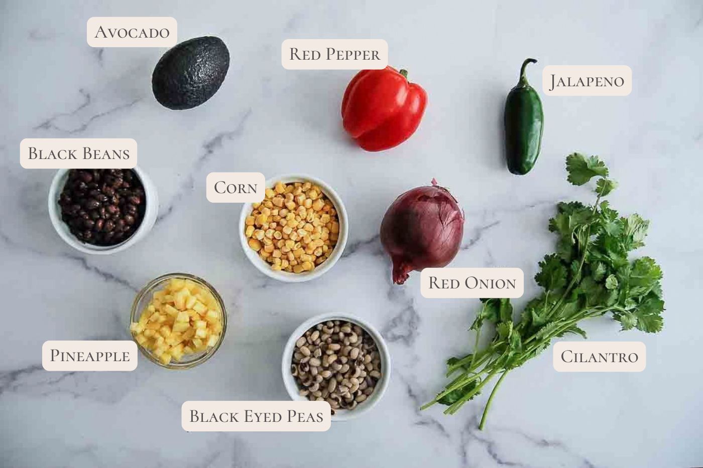 A marble surface displays labeled ingredients for texas caviar: avocado, red pepper, jalapeno, black beans, corn, red onion, pineapple, black-eyed peas, and cilantro, each positioned separately with text tags.