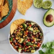 A bowl of texas caviar withbeans, corn, diced avocado, surrounded by tortilla chips, fresh cilantro, a halved avocado, and squeezed limes on a white surface.
