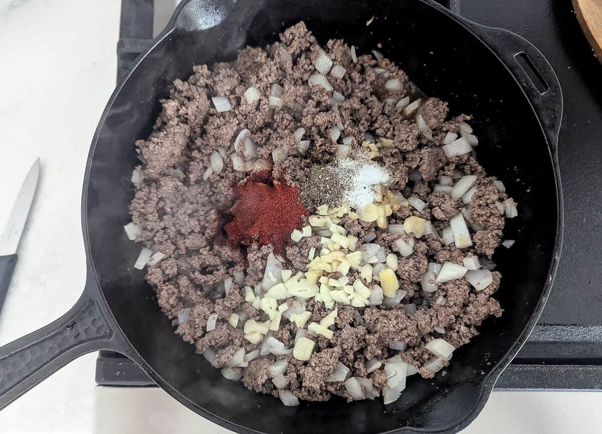 Ground beef cooking in a cast-iron skillet with chopped onions, minced garlic, paprika, salt, and pepper.