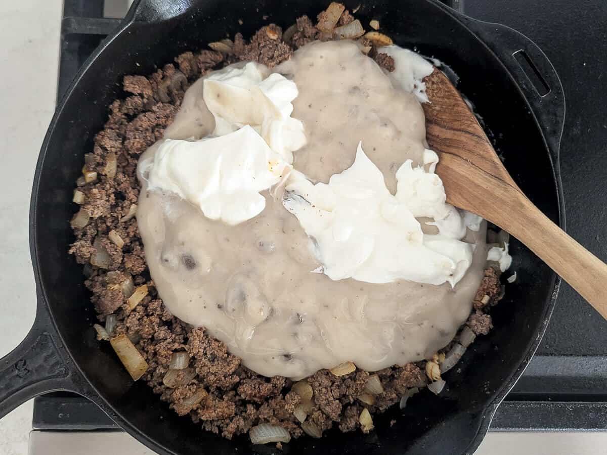 Ground beef and onions in a cast iron skillet topped with cream of mushroom soup and dollops of sour cream, with a wooden spoon resting on top.