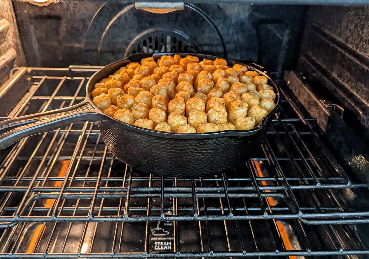 Cast iron skillet filled with a layer of golden-brown tater tots inside an oven.