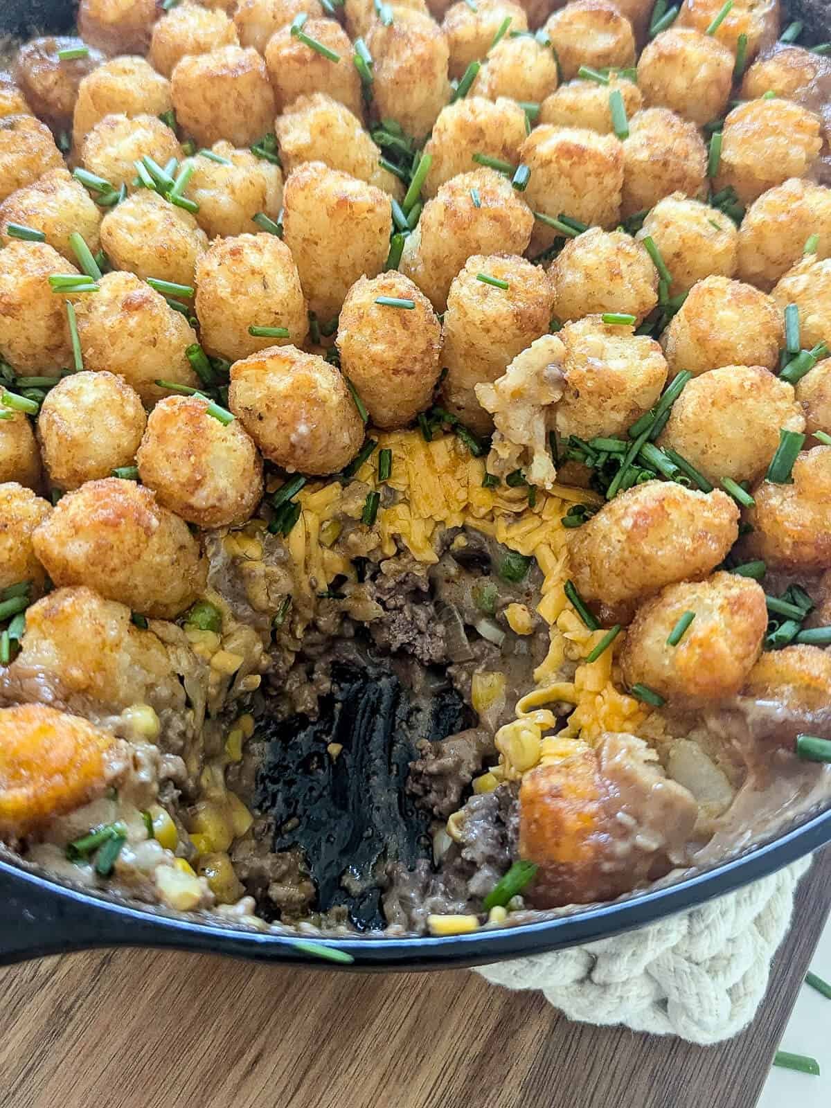 A cast iron skillet filled with tater tot casserole topped with chopped chives, with a portion scooped out to reveal a ground meat and vegetable filling.