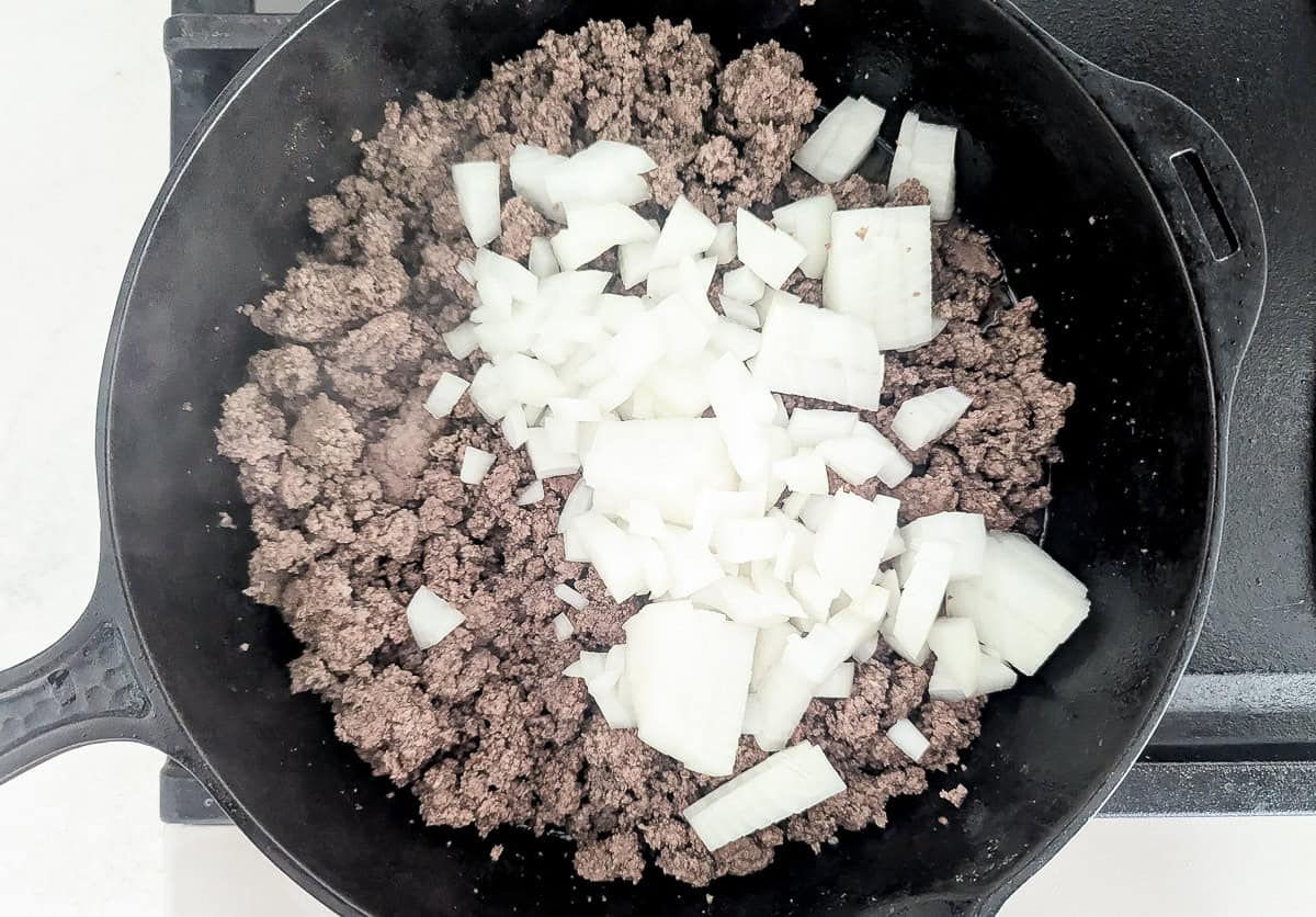 Ground beef browning in a cast-iron skillet with chopped white onions on top.